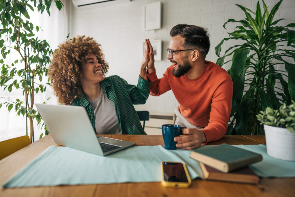 Two workmates excited about new job success\. High five\. Man and woman, colleagues, sitting at the table and working together using laptop\.