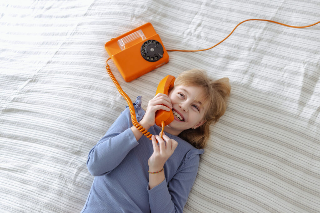 A cheerful girl lies on a striped bed, smiling while talking on an orange retro landline phone\. A cozy, nostalgic moment that suggests family, communication, and everyday joy\.